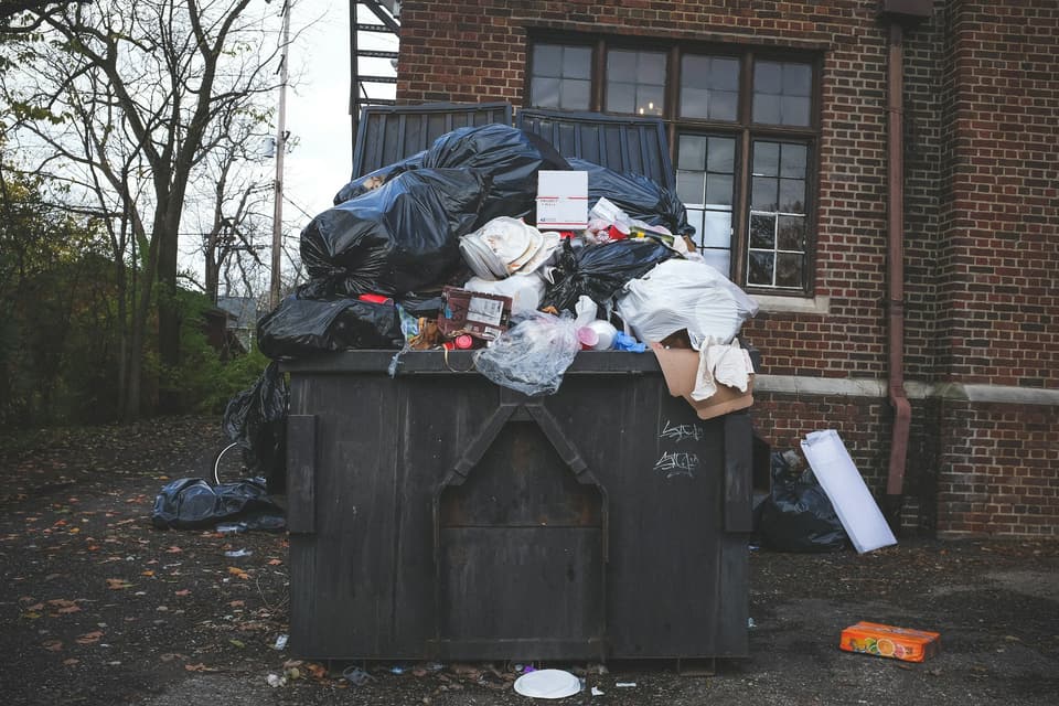 An overflowing trash dumpster piled with bags beside a brick building